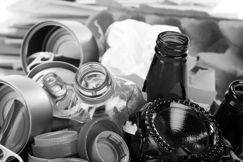 A close-up view of various discarded items in a rubbish collection scene, featuring several crumpled white paper, transparent glass bottles with black and brown tint, and multiple plastic bottle caps of different sizes and colours scattered across the foreground. In the background, there are larger items including a partially visible large plastic container, a crumpled paper bag, and other miscellaneous waste materials. The scene appears to be set outdoors, possibly on a pavement or driveway, with the waste arranged haphazardly as part of a rubbish removal process undertaken by Commercial Waste Removal Dalston, a service operating within the Dalston area. The amount of mixed waste materials suggests an ongoing effort to clear domestic or commercial rubbish, with the focus on recyclable plastics and glass bottles typical of general waste collection for disposal or recycling in London postcodes.