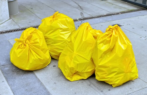 Four large yellow rubbish bags made of plastic material, with slightly crinkled textures, are placed on a paved concrete surface. The bags are loosely tied at the top, causing slight gathering of the material, and appear to be filled with waste. They are positioned in a cluster towards the left side of the image, with the largest bag at the back and other three bags in the foreground, slightly overlapping each other. The surrounding environment includes a section of grey paving slabs and a drainage grate in the background, indicative of an outdoor urban area, possibly a driveway or pavement in Dalston, London. The lighting suggests natural daylight. This scene is typical of waste collection or rubbish removal activities that companies like Commercial Waste Removal Dalston undertake in local residential or commercial settings to manage household or business refuse effectively.