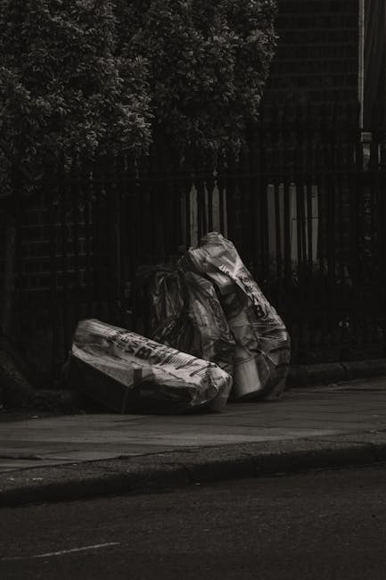 Three large black bin bags filled with rubbish are placed on the pavement next to a black metal fence, with a hedge and tree visible behind the fence. The bags appear to be made of plastic, with some