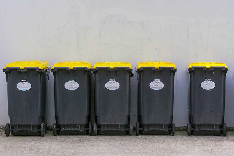Three large black bin bags filled with rubbish are placed on the pavement next to a black metal fence, with a hedge and tree visible behind the fence. The bags appear to be made of plastic, with some